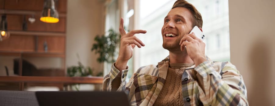 Lifestyle shot of young happy man sitting in cafe with laptop, answer phone call, talking to friend on mobile and laughing, chatting to someone with joyful face, resting in chair.