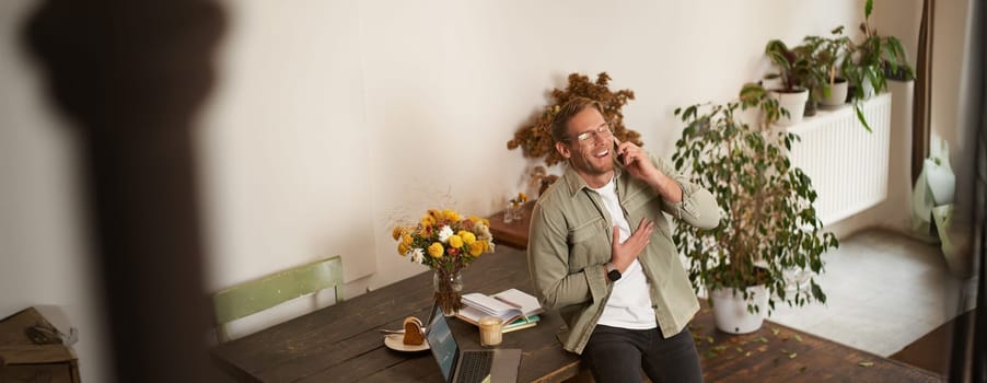 Portrait of handsome office manager, guy in co-working space, talking on mobile phone, calling someone, laughing and smiling, having pleasant conversation, sitting in office on table.