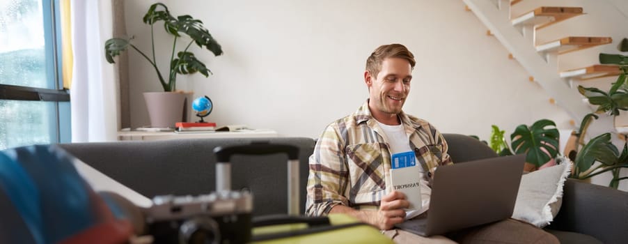 Portrait of handsome young man, has two tickets and passport, goes online on laptop, booking hotel and planning his holiday, sitting in living room.