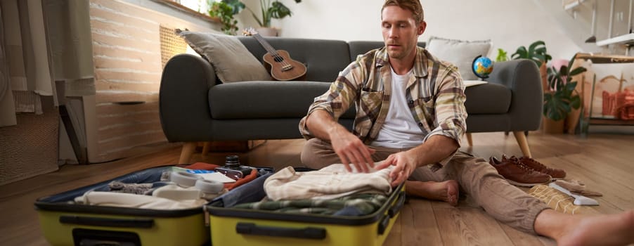 Image of young man, traveller, sitting on floor with opened suitcase, packing clothes for a trip, going on holiday. Tourism concept