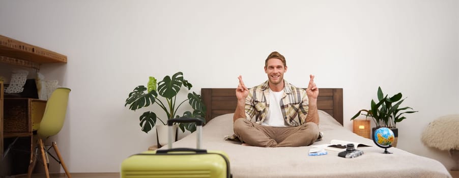 Portrait of young man meditating on a bed after packing suitcase for summer holiday, sitting with crossed legs in zen, nirvana pose, relaxing, hoping for something. Tourism and mindfulness concept