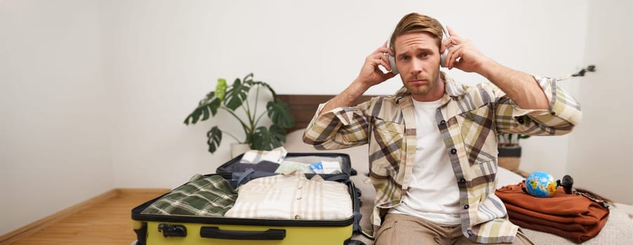 Portrait of handsome young man packing things in suitcase, listening to music in headphones, tourist going on vacation, preparing for holiday trip.