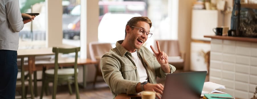 Happy smiling guy in glasses, sits in cafe, shows peace sign at laptop camera, video chats, connects to online meeting.
