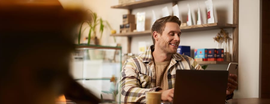Lifestyle portrait of young businessman, freelancer sitting in cafe, drinking coffee and using laptop, working on online project, digital nomad doing his task, has remote job, looking satisfied.