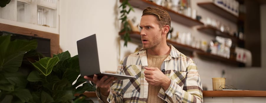 Portrait of man looking stressed and concerned at laptop screen, clench fist and frowns with angry face expression, sits in cafe with cup of coffee.
