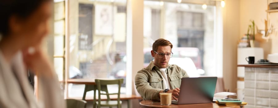 Image of young digital nomad, man in glasses sits in cafe, works from coffee shop, uses laptop in co-working space, wears glasses, drinks his beverage.