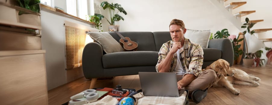 Image of young man, tourist going on holiday, sitting with a dog in living room, looking at laptop, check-in on a flight on computer. Tourism and travelling concept