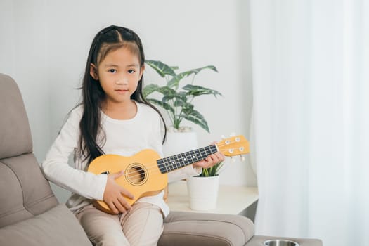 Portrait of a young girl in a white shirt holding a wooden ukulele on a grey couch. Her confident gaze at the camera adds a lively charm to the bright and modern living room.