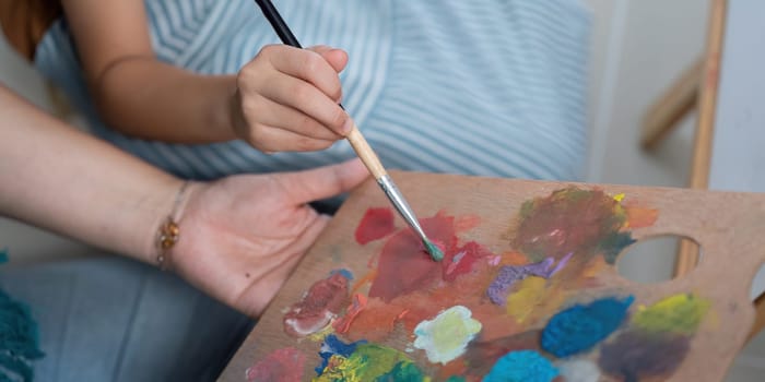 A daughter learns painting techniques from her mother, who guides her in selecting colors from a vibrant palette.