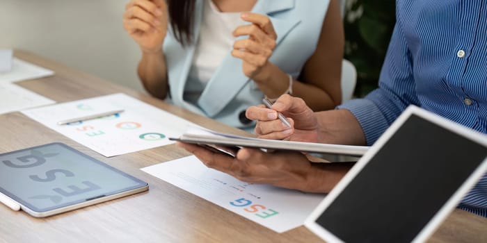 Two professionals collaborating in an office setting, analyzing ESG strategies on a tablet and papers, reflecting teamwork and sustainability efforts.