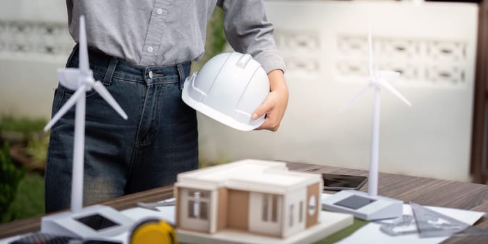An engineer holding a helmet examines a sustainable architectural model featuring wind turbines, showcasing innovation in eco-friendly building design.