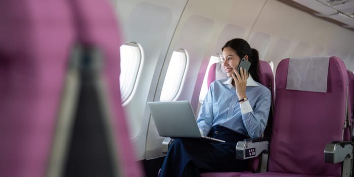 A professional Asian woman using a laptop and talking on the phone while seated on an airplane, embodying productivity and comfort during business travel.