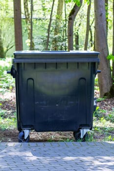 A large black trash can is currently stationed on a sidewalk that runs through the woods, providing a place for waste disposal