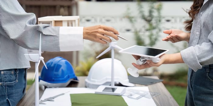 A sustainability engineer exchanging ideas with a colleague about renewable resources, showcasing building models and wind turbines in a collaborative outdoor environment.