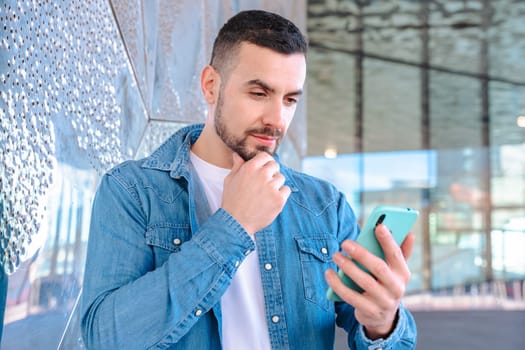 young man wearing a denim shirt, standing outdoors near a reflective glass wall, thoughtfully looking at his smartphone in natural daylight, symbolizing communication and modern life.