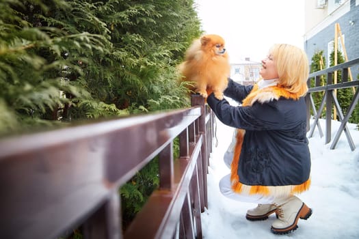 A middle-aged woman lovingly lifts her fluffy Pomeranian while enjoying a snowy winter day outdoors.