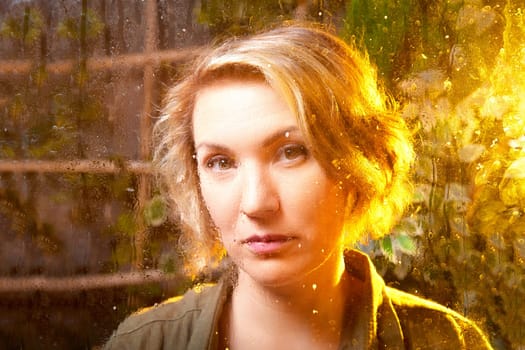 A woman with short blonde hair gazes thoughtfully through a misted window, surrounded by greenery, highlighting a tranquil atmosphere.