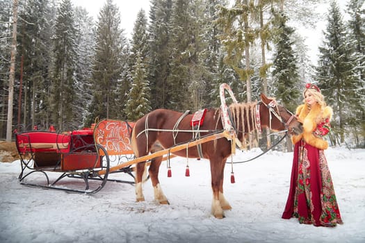 Russian girl in a vibrant national outfit poses with a horse and sled, celebrating Maslenitsa in winter.