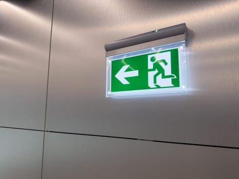 Emergency exit sign with left arrow and running man symbol mounted on metal wall. Green illuminated panel used for evacuation guidance in public building. Concept of safety, escape and compliance
