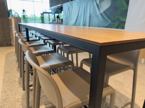 Long wooden high table with bar chairs in bright modern interior. Stylish cafeteria or coworking seating zone. Clean open space designed for comfort, dining, socializing and informal meetings