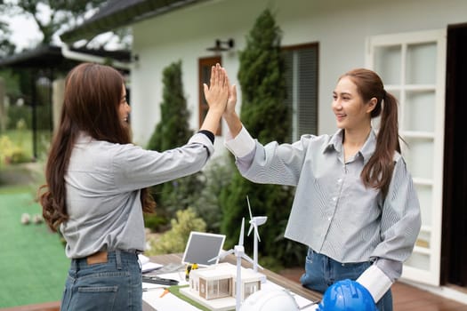 Two sustainability engineers share a high-five outdoors to celebrate their successful project, with blueprints and models displayed on their table.