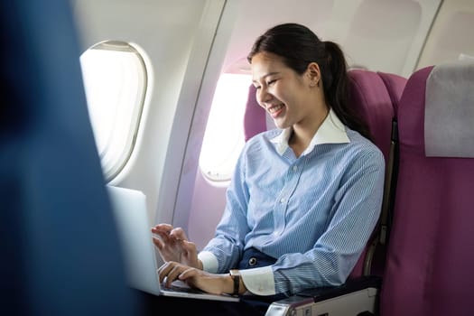 A cheerful young woman engages with her laptop on an airplane, enjoying her business travel while connected with work.