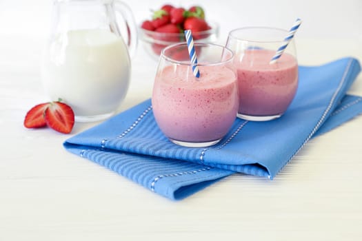 Two colorful strawberry smoothies in clear glasses are placed next to a pitcher of milk and a bowl of fresh strawberries on a blue napkin.
