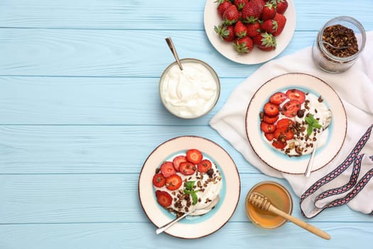 Two bowls filled with yogurt topped with sliced strawberries and granola sit on a light blue wooden table alongside honey, fresh strawberries, and utensils.
