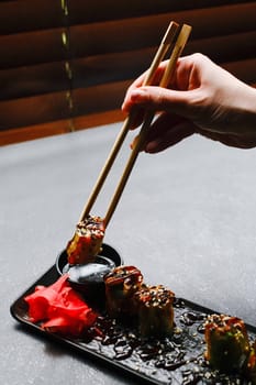 Hand Using Chopsticks to Pick Up Sushi on Black Plate with Ginger Garnish on Grey Table