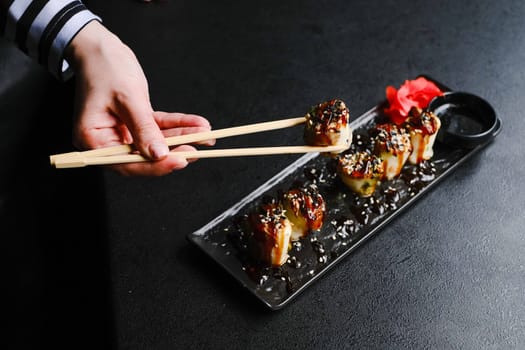 Hand Holding Chopsticks Picking Up Sushi Platter With Sauce On Black Table