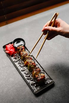 Hand Holding Chopsticks Picking Up Colorful Sushi Rolls on a Black Platter with Sauce