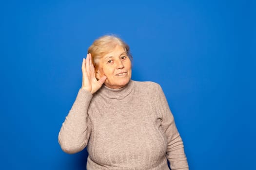 Caucasian woman in her 70s wearing a dark beige sweater putting her hand to her ear because she can't hear the most interesting gossip, isolated on blue background.