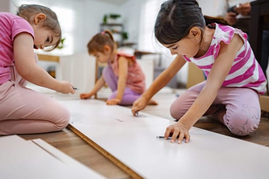 Three young girls gather on a wooden floor, focused and engaged in a fun crafting activity. They use pencils to draw on large sheets of paper, showcasing their artistic flair.