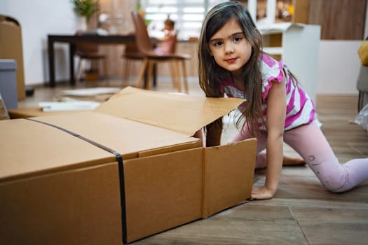 In a cozy living room, a young girl with long hair smiles as she peeks out from a large cardboard box. Excitement fills the space as her family is engaged in unpacking and organizing.