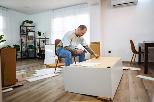 In a cozy, well-lit room, a focused man is piecing together furniture. Surrounded by packaging materials, he meticulously examines each component, creating a welcoming atmosphere.