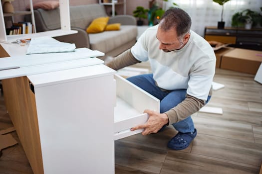 A man focuses intently as he fits a drawer into a newly built piece of furniture in his stylish living room. Surrounding boxes and tools hint at a productive day of assembling.
