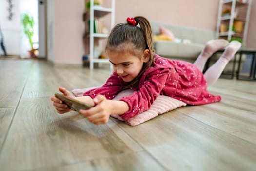 A young girl in a bright, cozy room keeps herself entertained by intensely focusing on her smartphone.