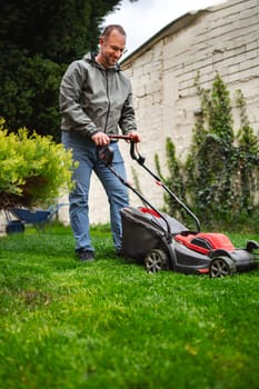 A man mows his vibrant green lawn with an electric mower, enjoying a sunny weekend afternoon. The garden features neatly trimmed hedges and lush grass, reflecting a dedication to outdoor upkeep.