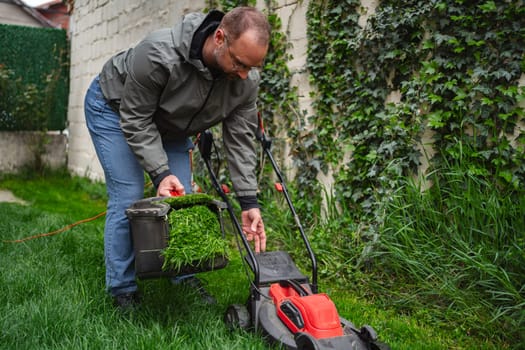 Gardener focuses on maintaining his vibrant lawn, emptying freshly cut grass clippings from a mower into a bucket. The surroundings are lush, highlighting a peaceful evening atmosphere.