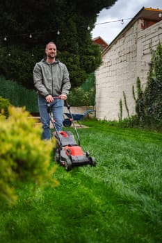 A man is carefully mowing his vibrant green lawn, showcasing neat lines across the grass. The garden is surrounded by a hedge and tall trees, creating a serene suburban atmosphere.