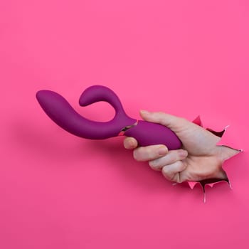 Female hand sticking out with raspberry sex toy from pink paper background