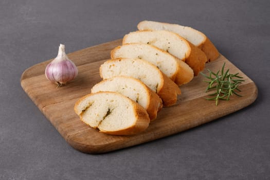 Freshly sliced bread with garlic and herbs displayed on a wooden cutting board on grey table.