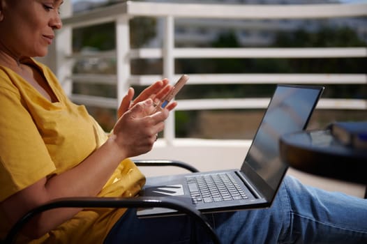 A woman sitting outdoors on a patio, using a smartphone and a laptop. She seems focused on her activity, enjoying a peaceful and productive moment in a comfortable setting.