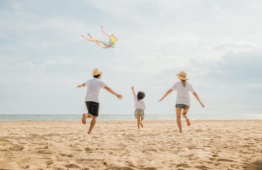 Asian family is flying a kite high in the sky while running on the beach. Happy parents mother and father with their child playing with kite, family is enjoying their time together, summer day
