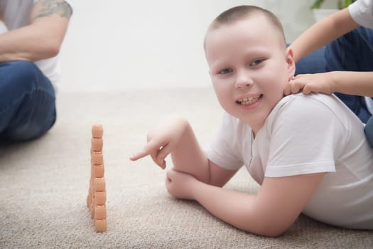 A young boy smiles as he stacks wooden blocks on the carpet with adults nearby, creating a fun playtime moment in a cozy environment.