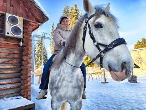 A person enjoys a horse ride atop a stunning white horse, surrounded by a serene winter landscape featuring snow-covered ground and a rustic wooden cabin.