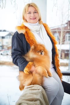 A blonde woman dressed warmly holds her small red Pomeranian while standing outside in the snow during winter.