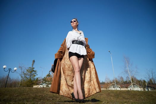 A woman stands confidently in a park, dressed in a long coat, high heels, and shorts, exuding a bold fashion sense under bright sunlight.