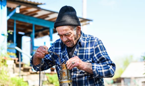 Elderly Russian man in a rural village holding a drainage pump with both hands, checking equipment under the open sky—simple village routine in retirement
