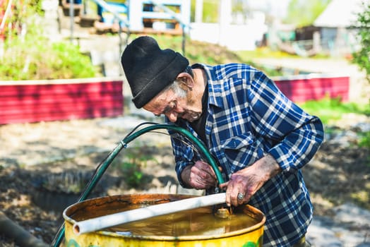 Elderly Russian man in a village working with a drainage pump, carefully inserting it into a barrel—retired but still active and engaged in rural tasks
